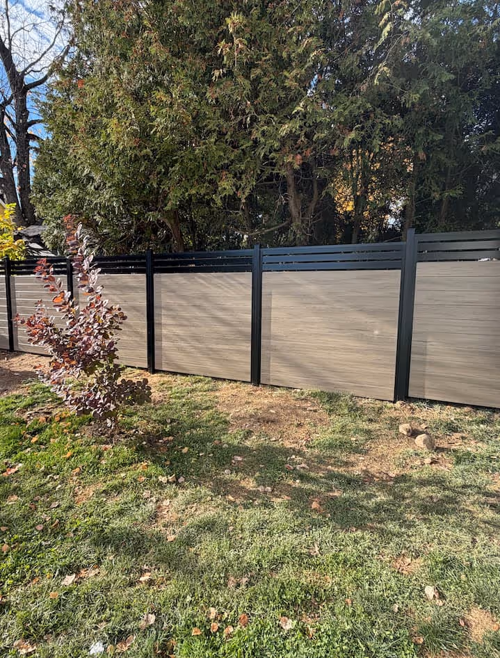 Clôture moderne avec panneaux taupe et cadres noirs devant des arbres verdoyants dans une cour herbeuse.