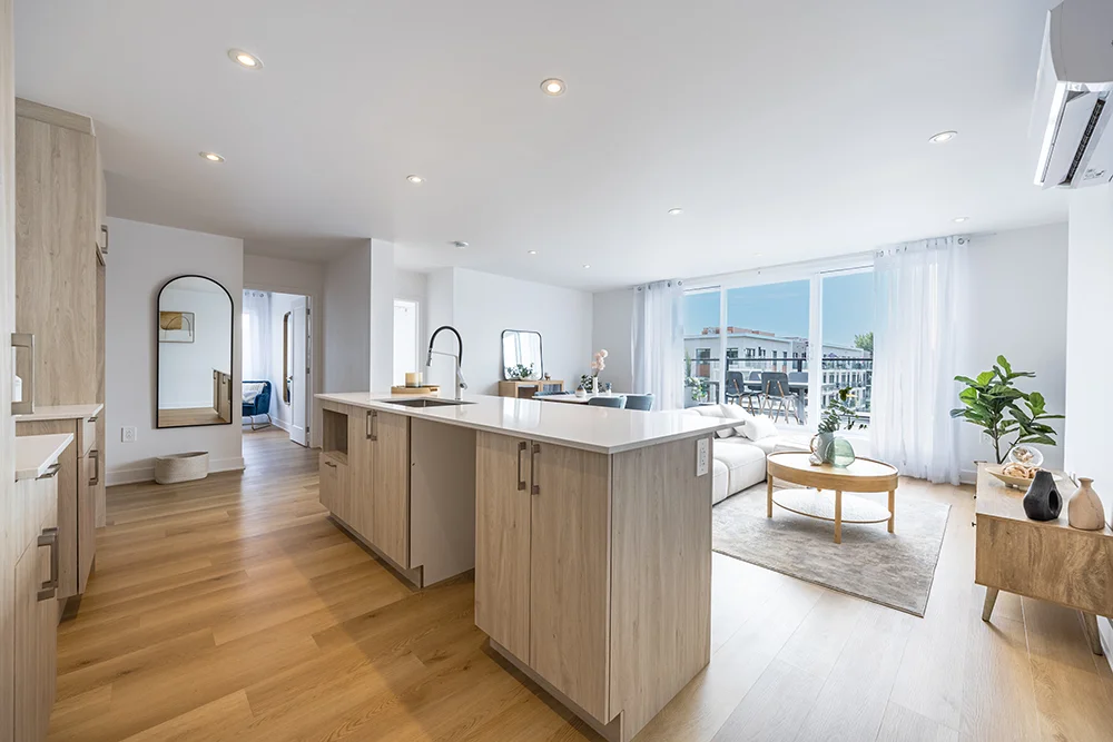 Modern open-concept kitchen and living room with wooden flooring, light wood cabinetry, large island, white sofa, and floor-to-ceiling windows opening to a balcony.