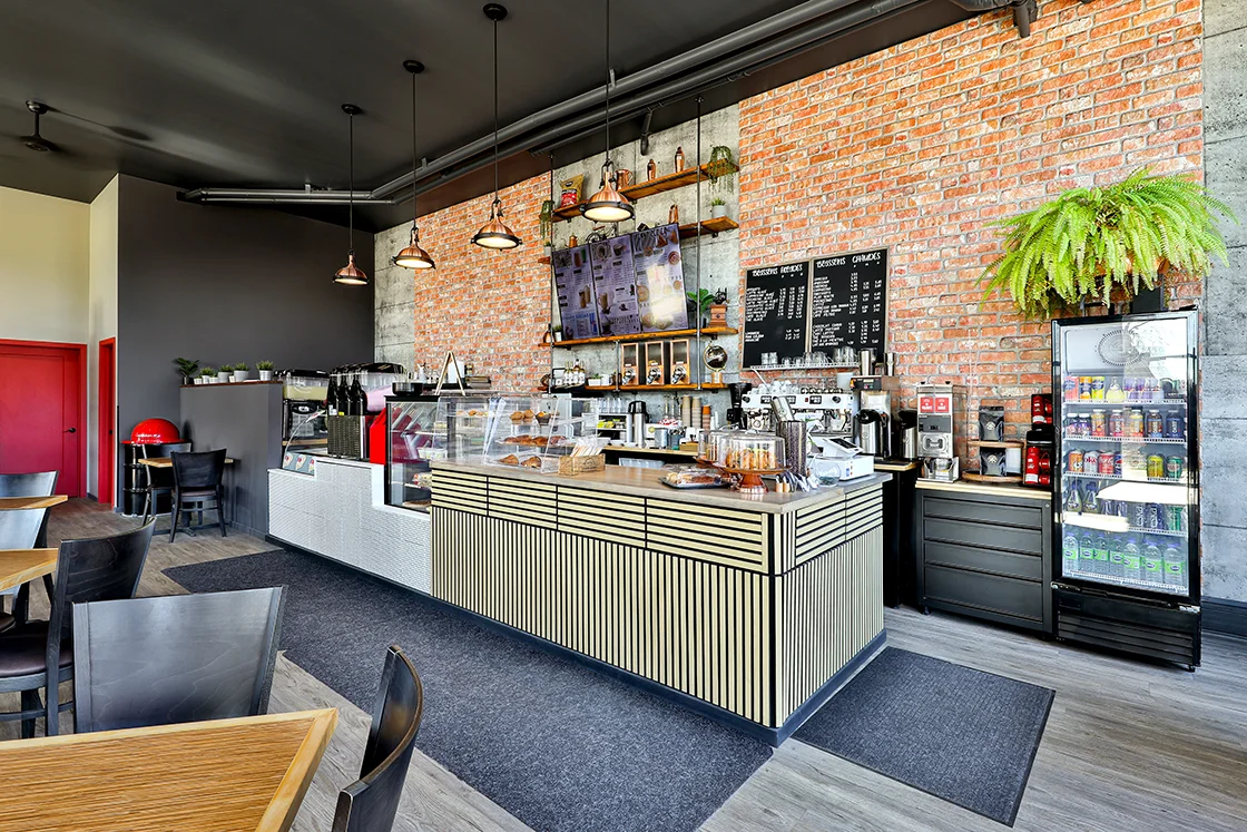 Modern cafe interior with wooden tables, black chairs, a striped counter displaying pastries, brick accent wall, menu boards, hanging lights, and a refrigerator stocked with drinks.