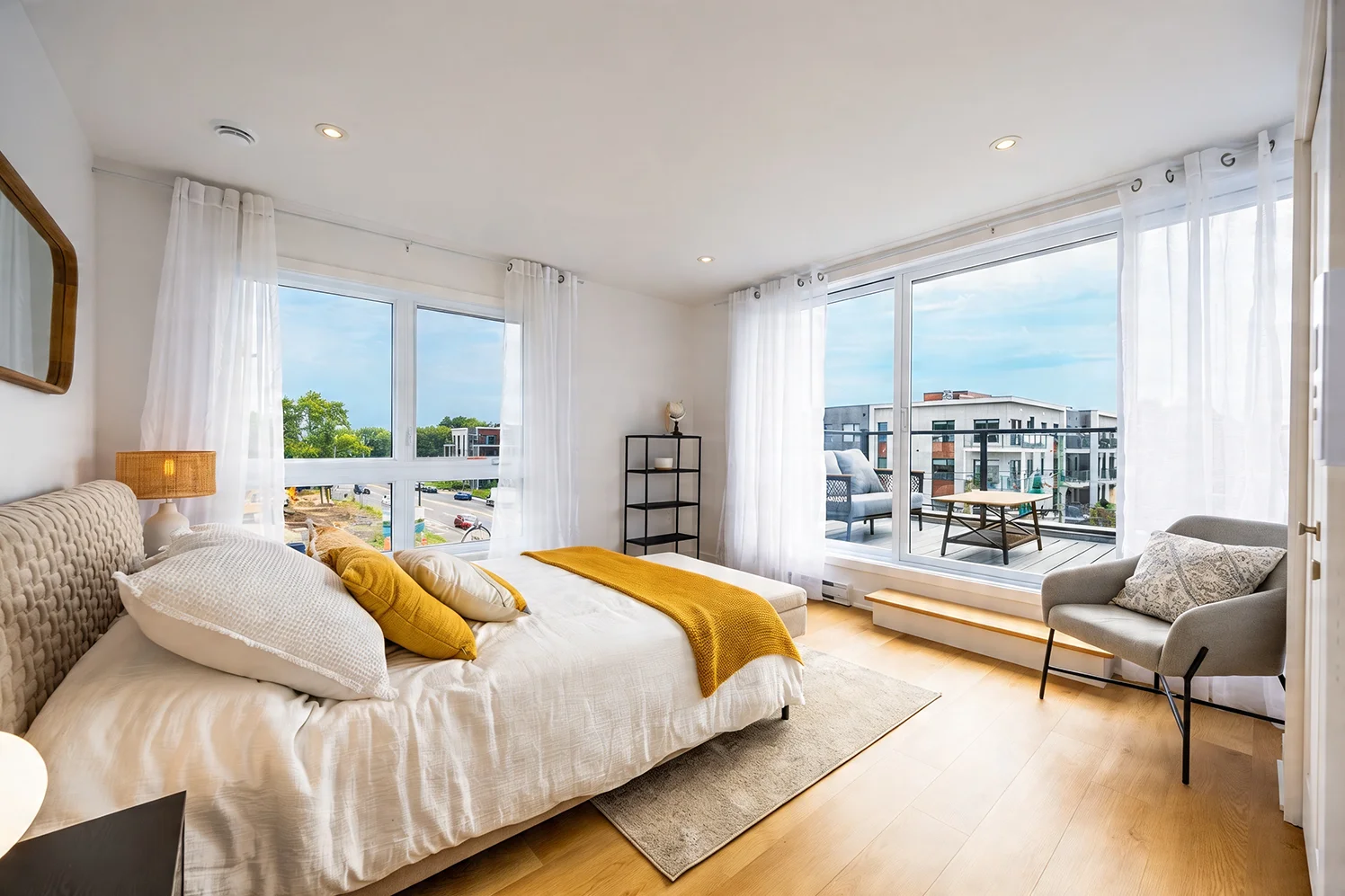 Bright modern bedroom with white bedding, mustard yellow pillows and throw, large windows with sheer curtains, a gray armchair, and a balcony view.