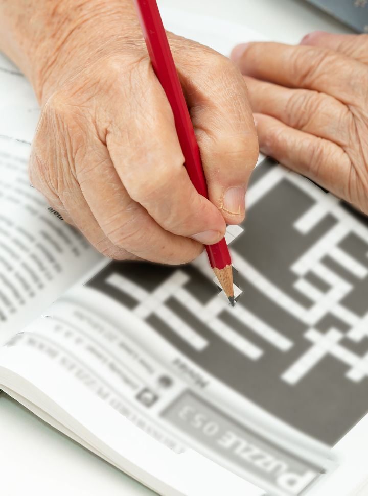 Close-up of an older adult's hands holding a red pencil while solving a crossword puzzle