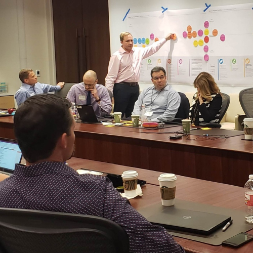 A man in a pink shirt points to a large chart with colorful sticky notes while four colleagues sit around a conference table, listening.