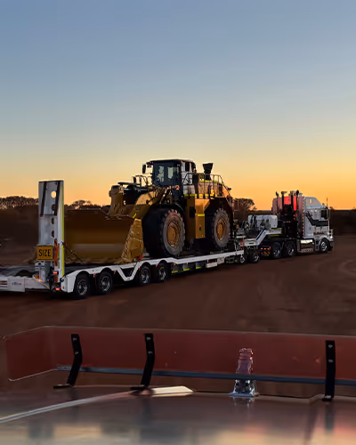 Yellow haul truck on a flatbed trailer at sunrise under a pink sky
