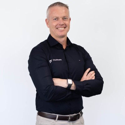 Smiling man with short gray hair wearing a black shirt with FlexWeblabs logo, arms crossed, standing against a white background.