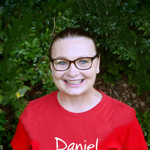 Smiling woman with glasses and hair pulled back wearing a red shirt standing in front of leafy greenery.