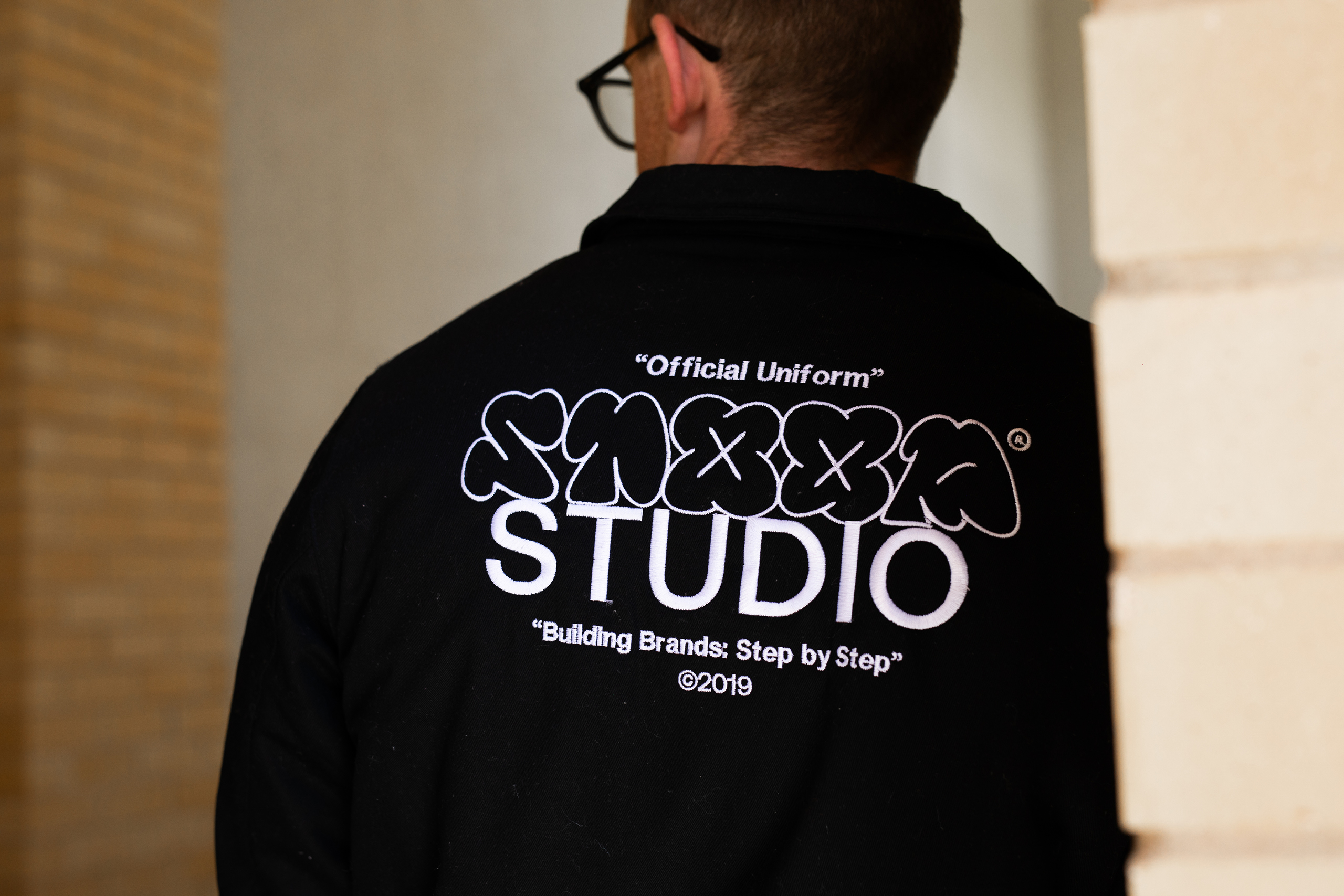Person wearing a black jacket with white text on the back reading 'Official Uniform' STOOP STUDIO 'Building Brands: Step by Step' ©2019.