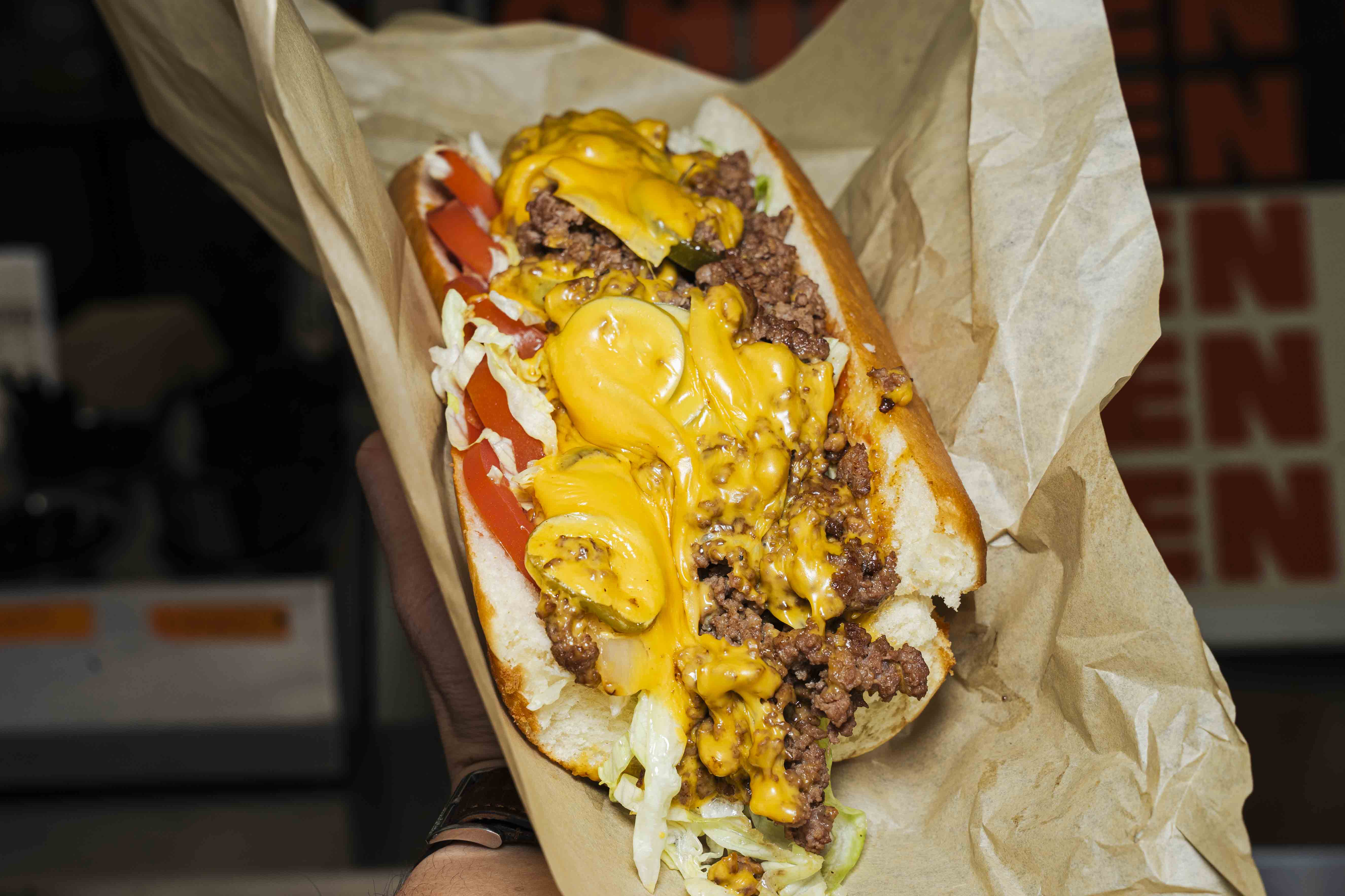 A hand holding a juicy chopped cheese sandwich loaded with ground beef, melted cheddar, lettuce, tomato, and jalapenos on a toasted hoagie roll.