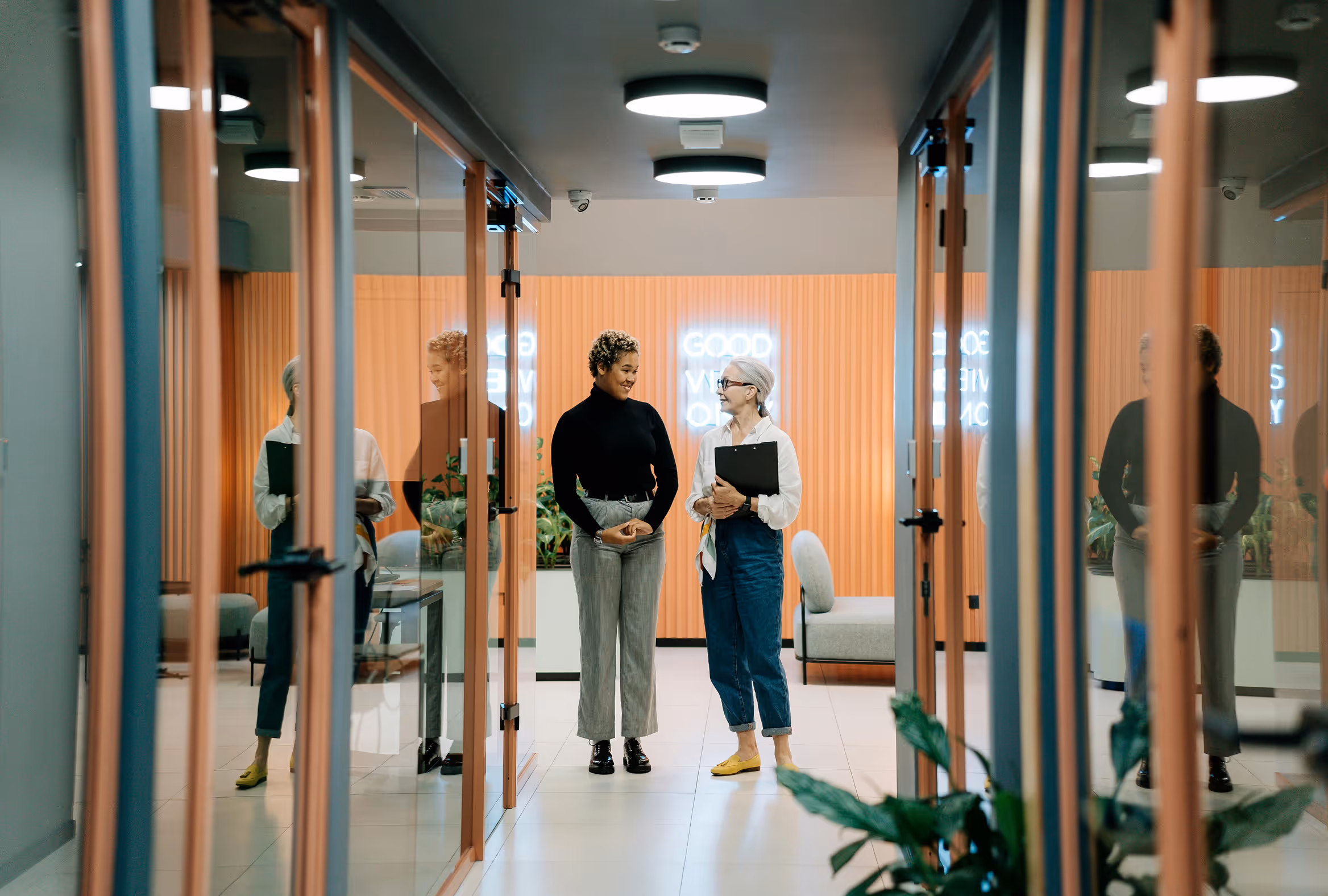 Two women conversing in a modern office hallway with glass doors and warm lighting. One is holding a tablet, the other a notebook.