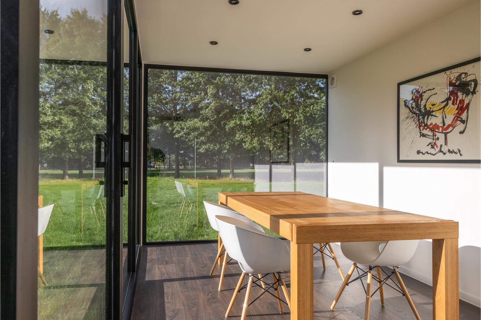 Een moderne eethoek met grote glazen wanden, een houten tafel, witte stoelen en ingelijste abstracte kunst aan de muur. Deze door ROBCOR ontworpen ruimte kijkt uit over een groen buitenlandschap en is voorzien van een multifunctionele unit en overvloedig zonlicht voor een open sfeer.
