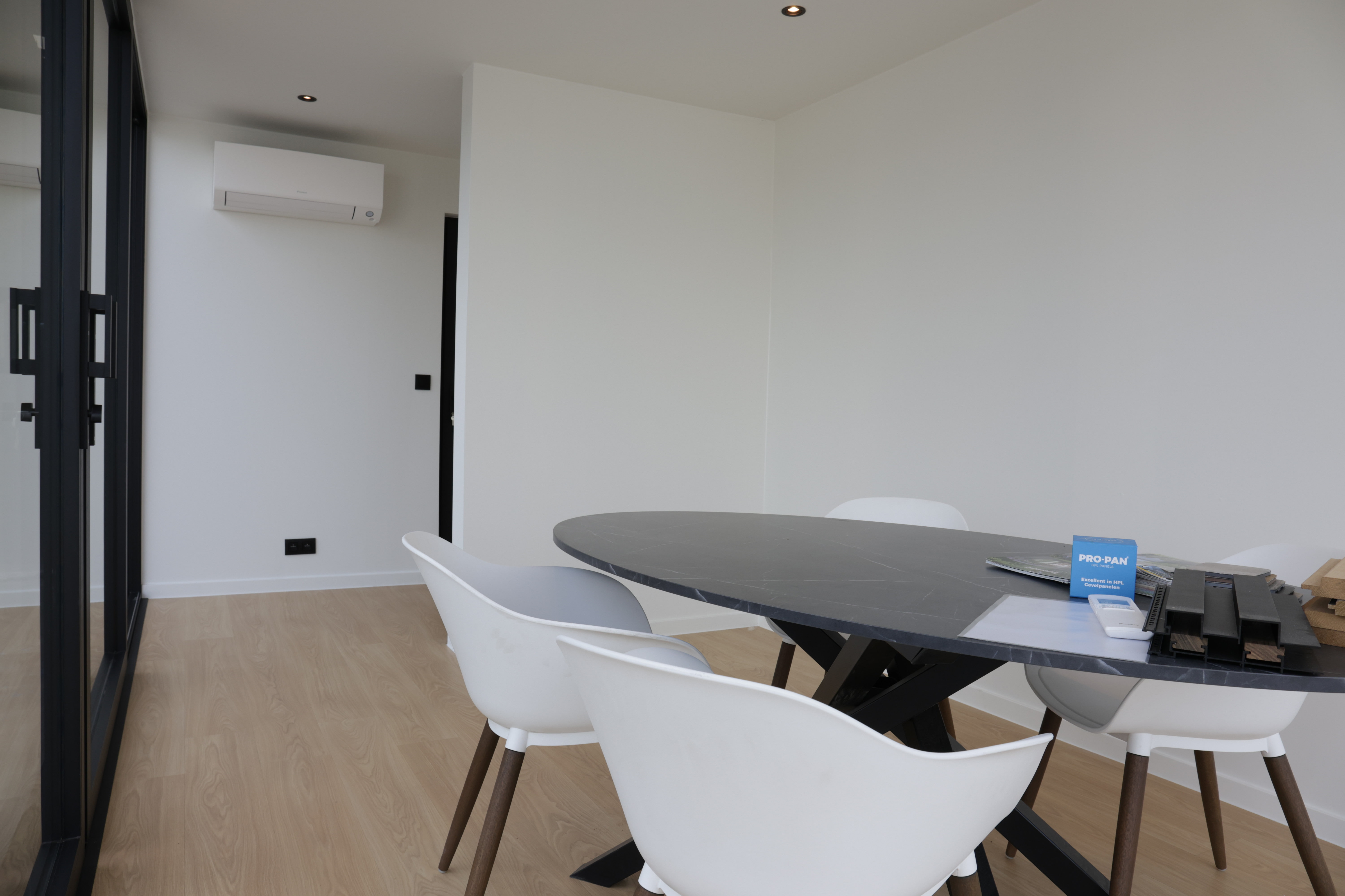 Een modern bureau met een ronde tafel en vier stoelen, gericht naar een grote glazen muur die een visueel verband legt tussen binnen- en buitenruimte. De overzichtelijke indeling en heldere kleuren creëren een rustige en functionele omgeving.