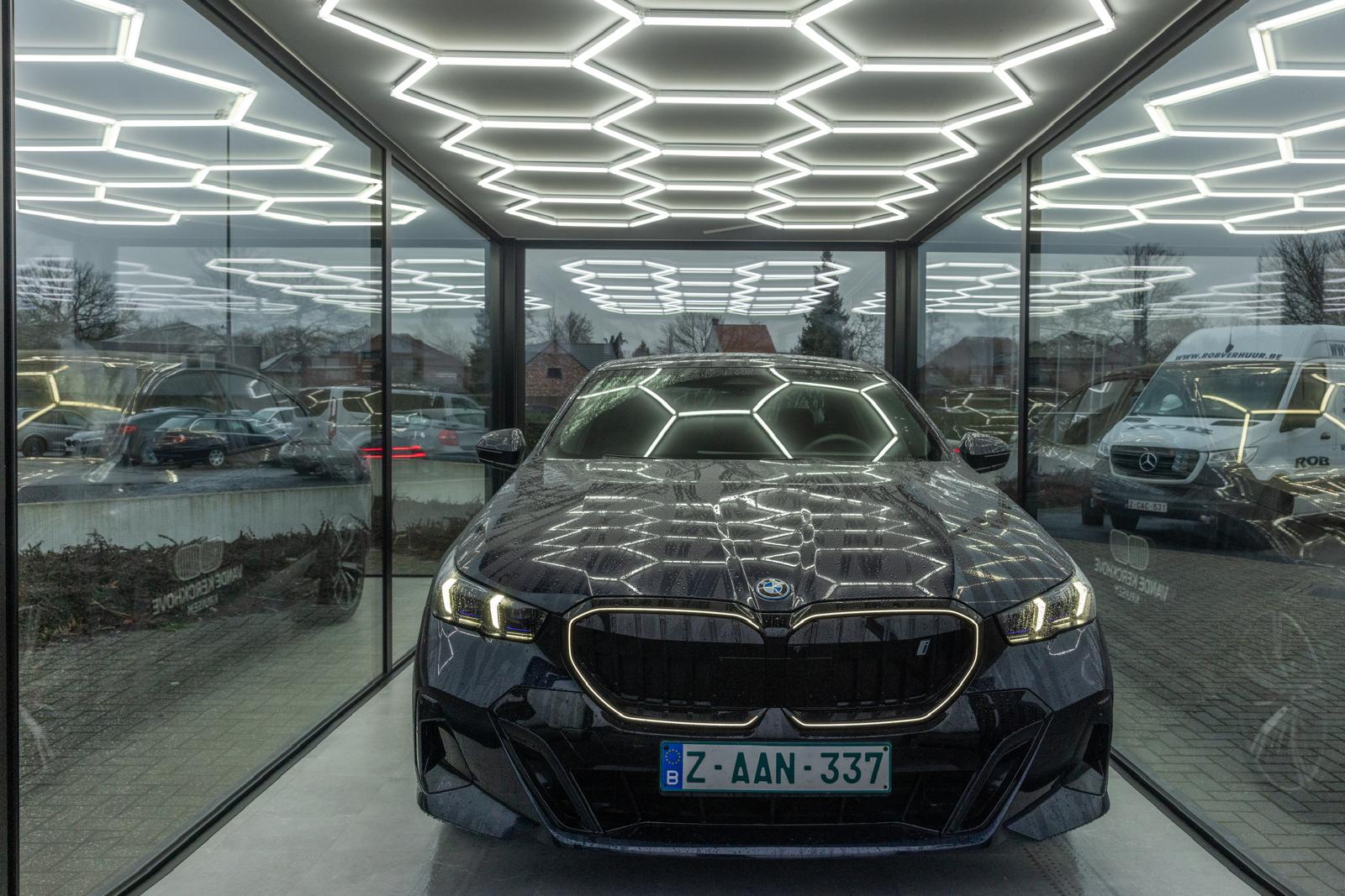 Een BMW-voertuig staat binnen in een glazen unit met hexagonale LED-verlichting aan de plafond. De auto is afgewerkt en heeft een modern ontwerp.