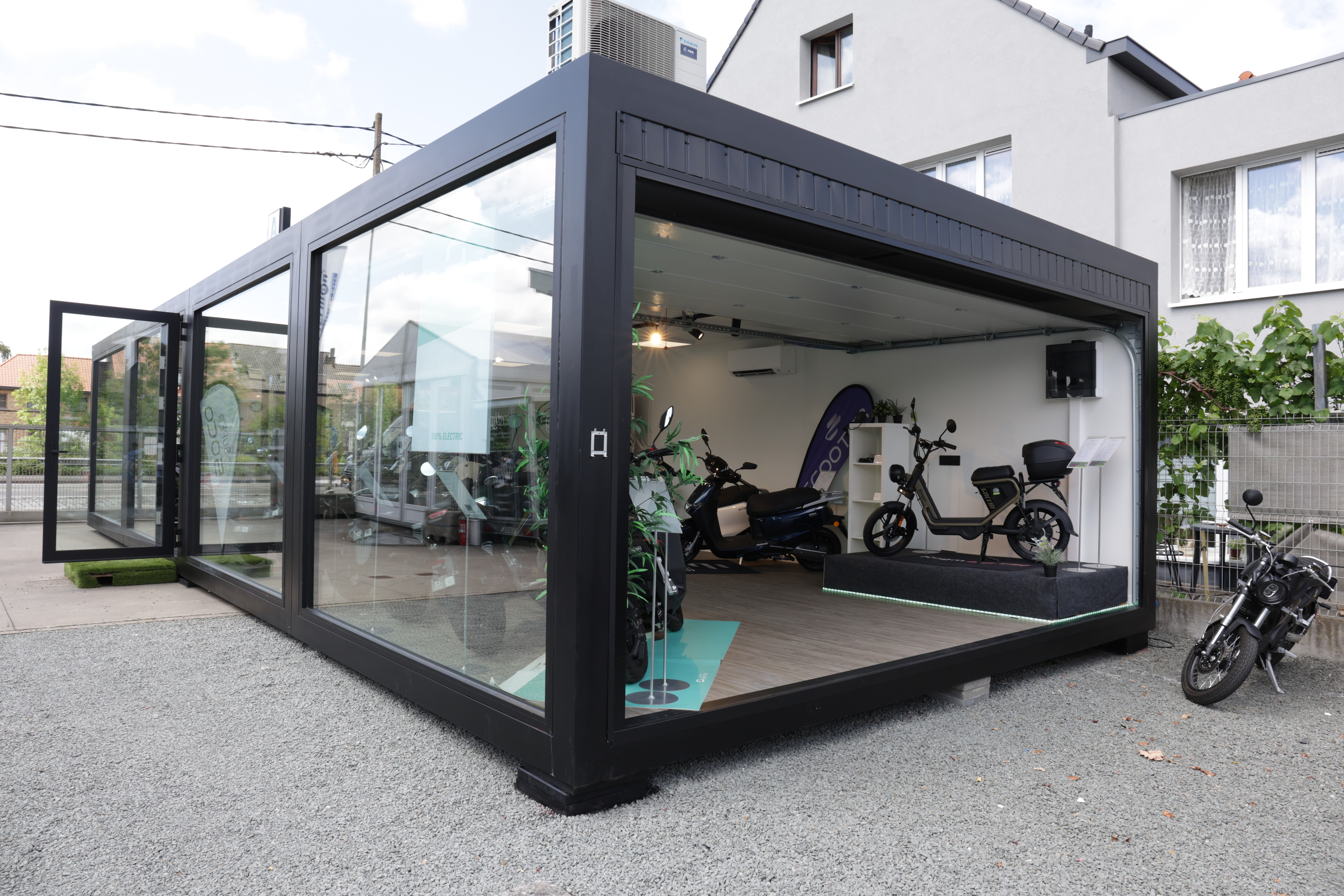 Moderne showroom in unit met grote glazen wanden, een open deur en een aan de muur gemonteerd airconditioningsysteem van ROBCOR. Binnen staan meerdere elektrische brommers tentoongesteld op een houten vloer, geschikt als presentatie- en verkoopruimte.