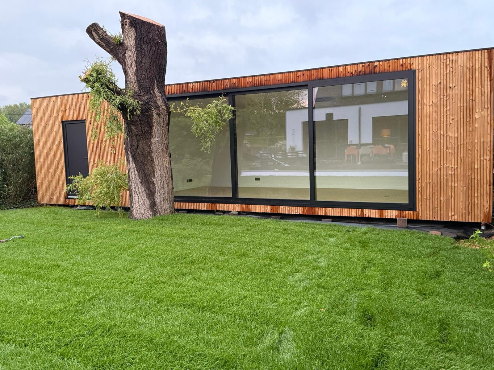 Een modern houten woonunit met glazen wanden, gelegen naast een grasveld en een struik, reflecteert een harmonieuze combinatie van natuur en architectuur.