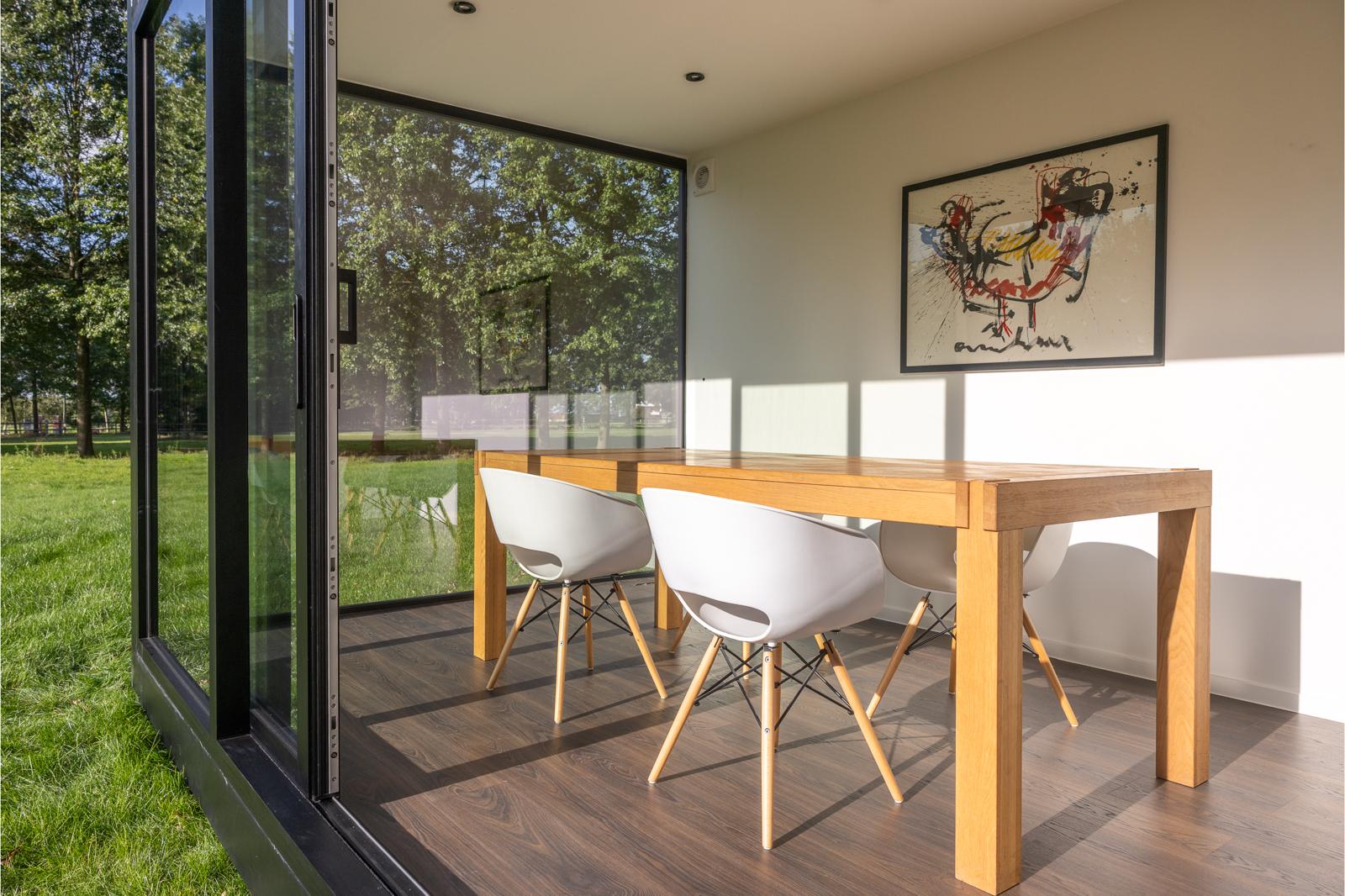 Een moderne eethoek met grote glazen wanden, een houten tafel, witte stoelen en ingelijste abstracte kunst aan de muur. Deze door ROBCOR ontworpen ruimte kijkt uit over een groen buitenlandschap en is voorzien van een multifunctionele unit en overvloedig zonlicht voor een open sfeer.