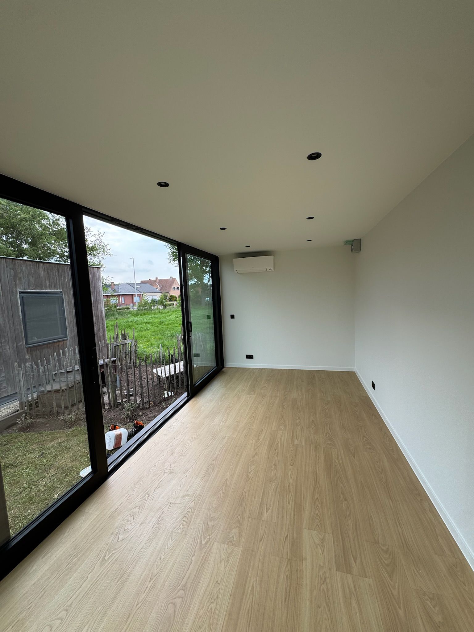 Moderne, lege ruimte met houten vloer, inbouwverlichting en groot raam met uitzicht op een grasrijke achtertuin – strakke en lichte interieurafwerking door ROBCOR.