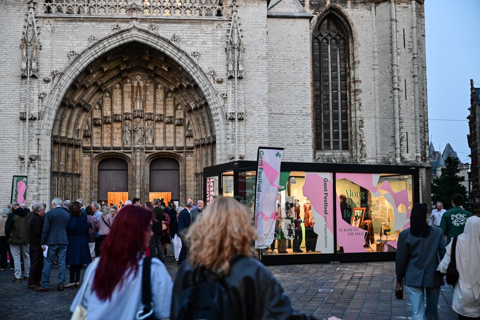 Een levendige straatopstelling met een tentoonstellingsstand voor "Gent Festival van Vlaanderen" naast een middeleeuwse kerk, waar mensen doorheen lopen en kijken naar de modulaire unit.