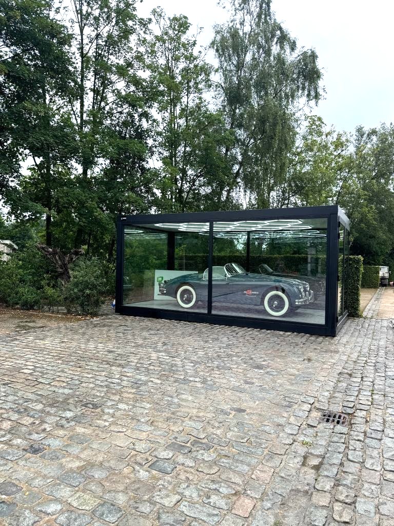 Klassieke zwarte auto tentoongesteld in een strak glazen unit op een kasseienoprit, omringd door weelderige groene bomen – straalt elegantie en nostalgie uit.