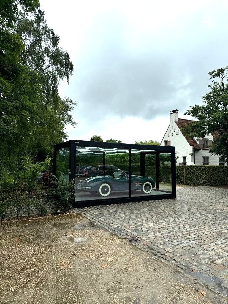 Klassieke zwarte auto tentoongesteld in een strak glazen unit op een kasseienoprit, omringd door weelderige groene bomen – straalt elegantie en nostalgie uit.