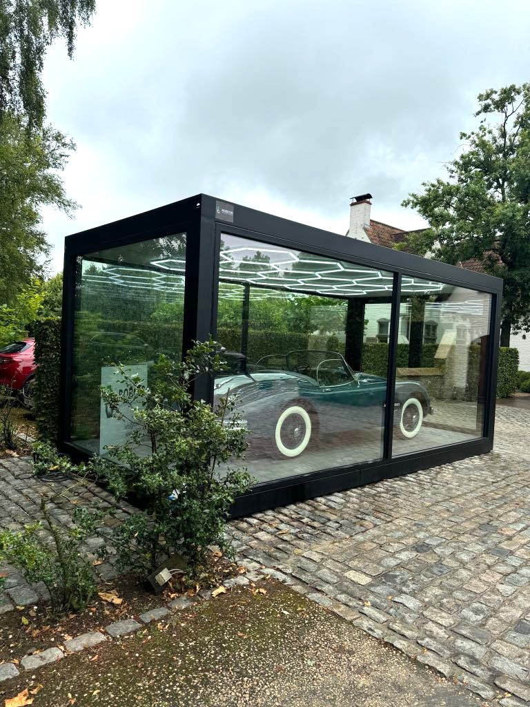 Klassieke zwarte auto tentoongesteld in een strak glazen unit op een kasseienoprit, omringd door weelderige groene bomen – straalt elegantie en nostalgie uit. 
