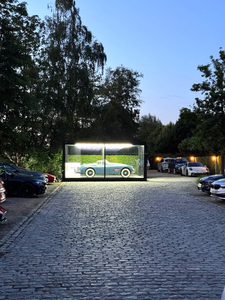 Vintage blauwe auto tentoongesteld in een goed verlichte glazen unit op een kasseienparkeerplaats, omringd door bomen bij schemering – sfeervol en rustig.