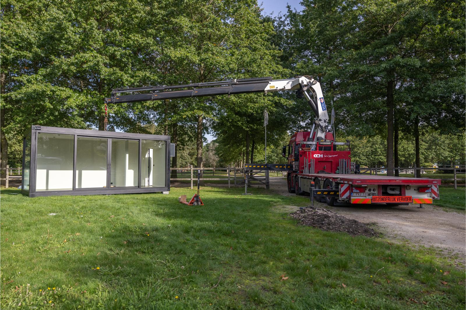 Rode kraanwagen hijst een modulaire unit op een grasrijke locatie omringd door bomen – professionele plaatsing of verplaatsing door ROBCOR in een rustige omgeving.