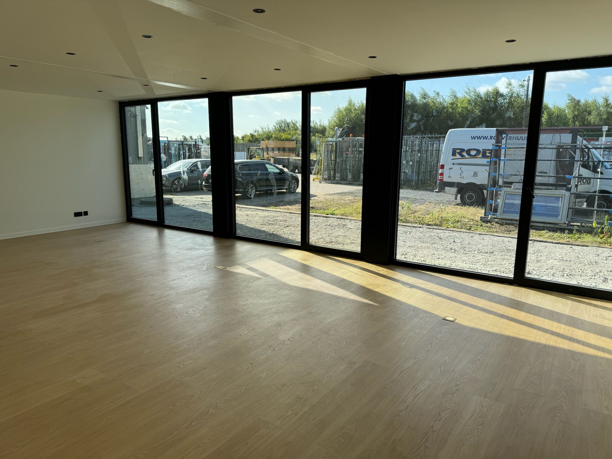 Ruime, lichte kantoorunit van ROBCOR met lichte houten vloer en grote raampartijen van vloer tot plafond, met uitzicht op een parkeerterrein buiten.