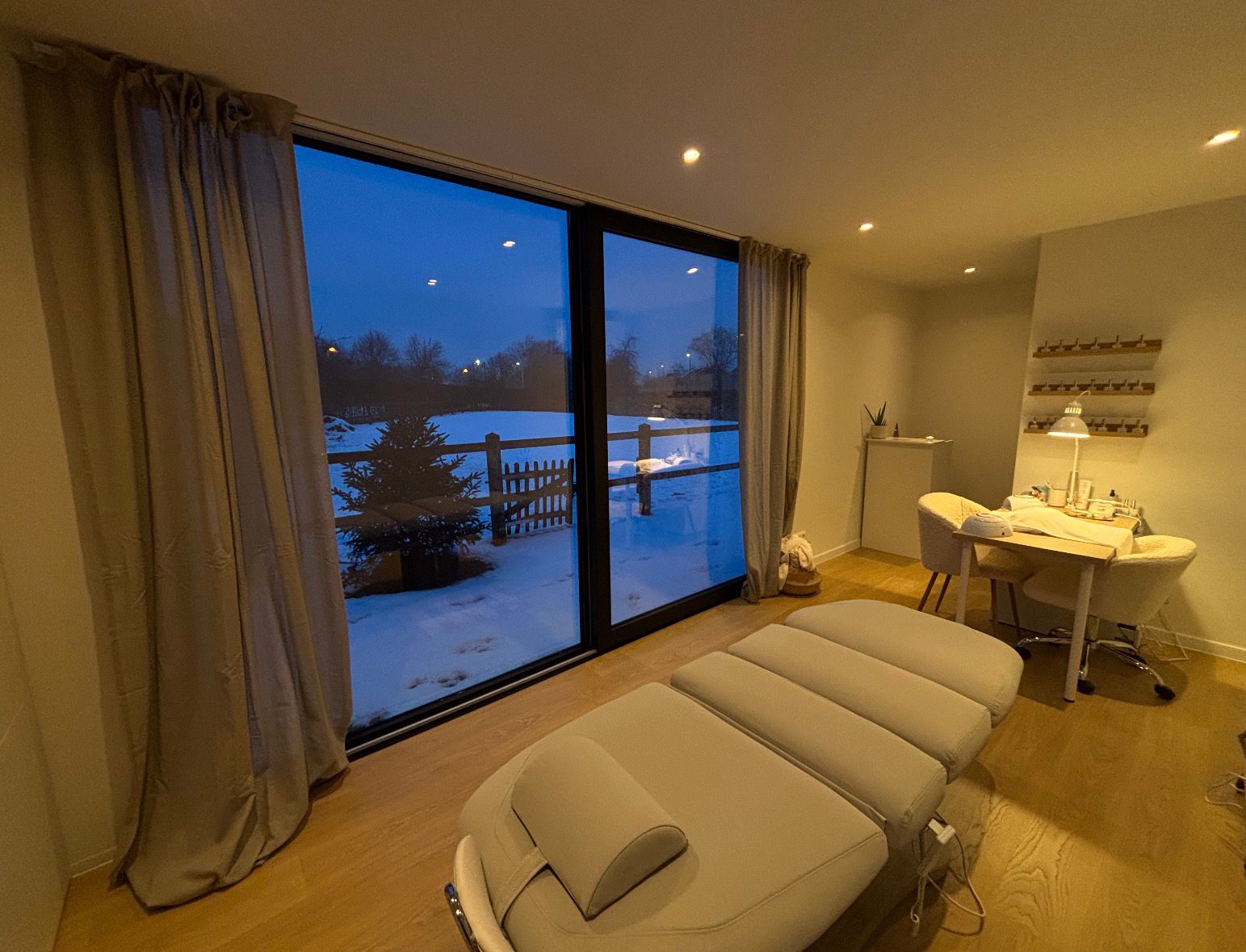 Modern beautysalon met beige behandelstoel, tafel met twee stoelen en grote ramen met uitzicht op besneeuwd landschap.