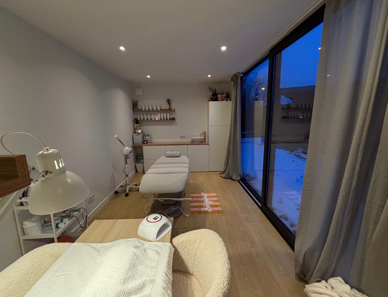 Rustige schoonheidssalon met behandeltafel, witte meubels, houten vloer en grote ramen met uitzicht op sneeuw buiten.