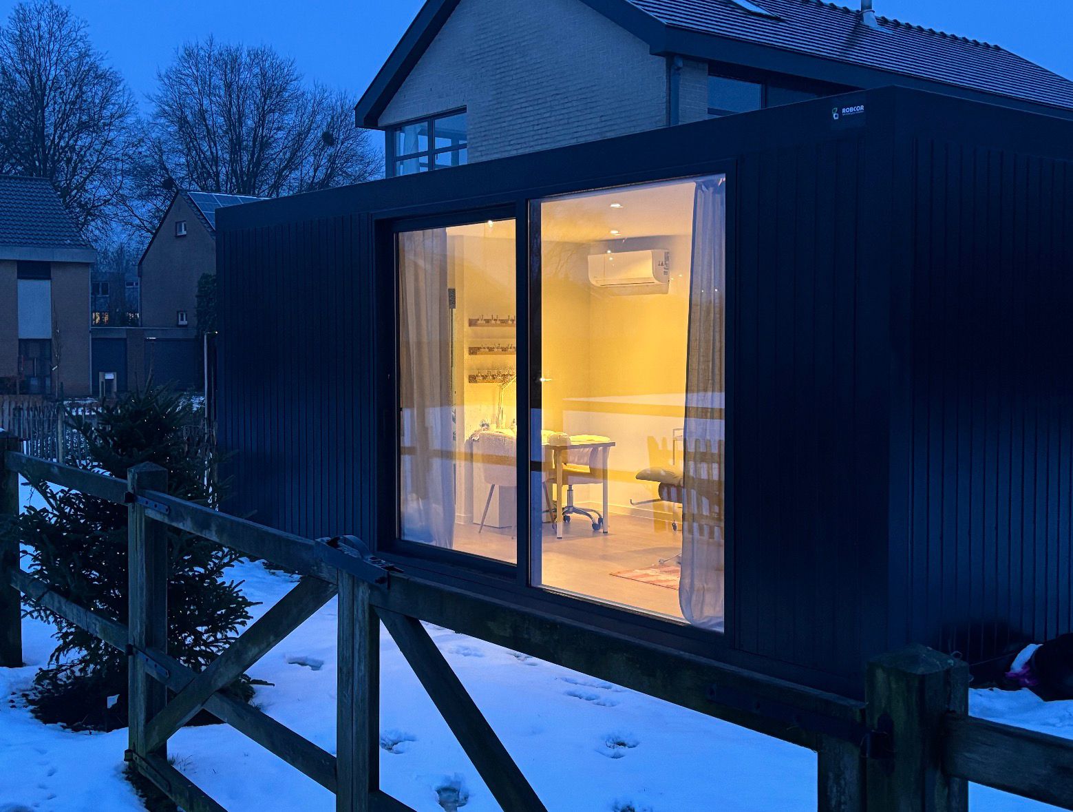 Verlichte moderne tuinstudio met grote glazendeur in een besneeuwde tuin bij schemering.