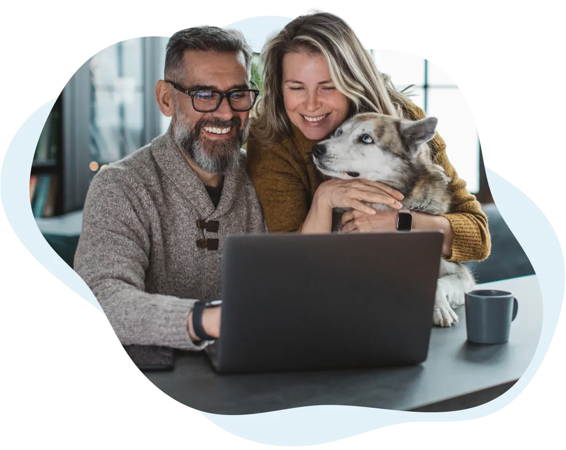 Smiling couple sitting together with their dog, looking at a laptop screen, in a cozy home setting.