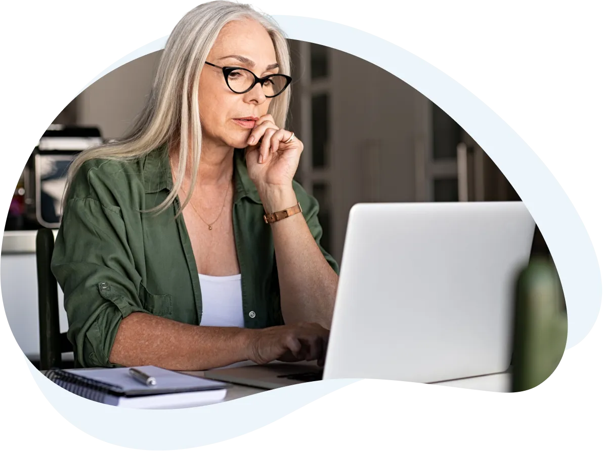 Older woman with long gray hair wearing glasses and a green shirt, thoughtfully working on a laptop at home.