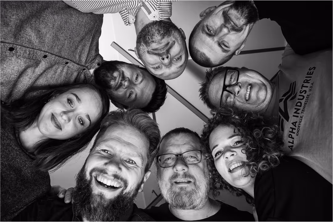 Žmonių grupė žiūri į fotoaparatą apskritimu, juodu baltame portrete.