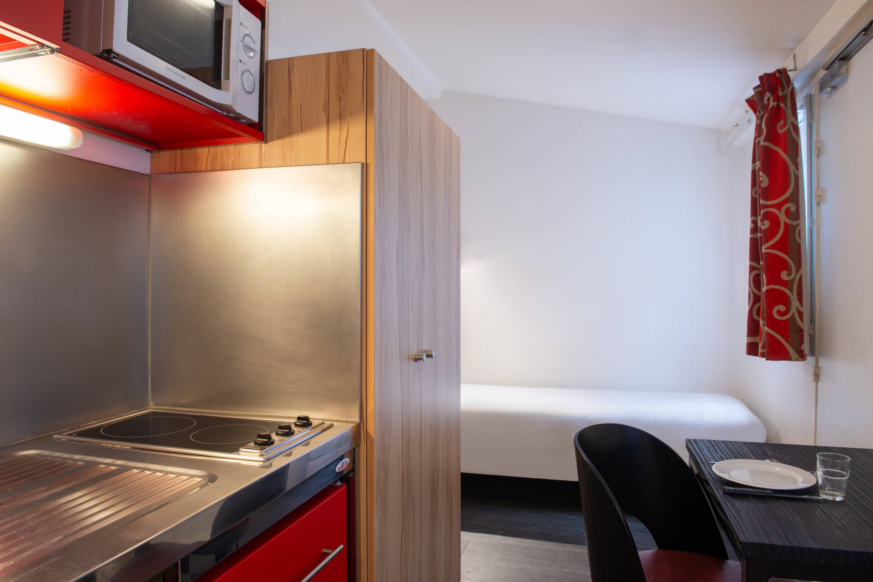 Image de studio de 10 m² pour 1 personne avec kitchenette rouge et acier avec frigidaire et micro-onde. Ce studio contient aussi une salle de bain blanche avec douche, lavabo, miroir et toilettes. Il y a un lit individuel et une télévision.
