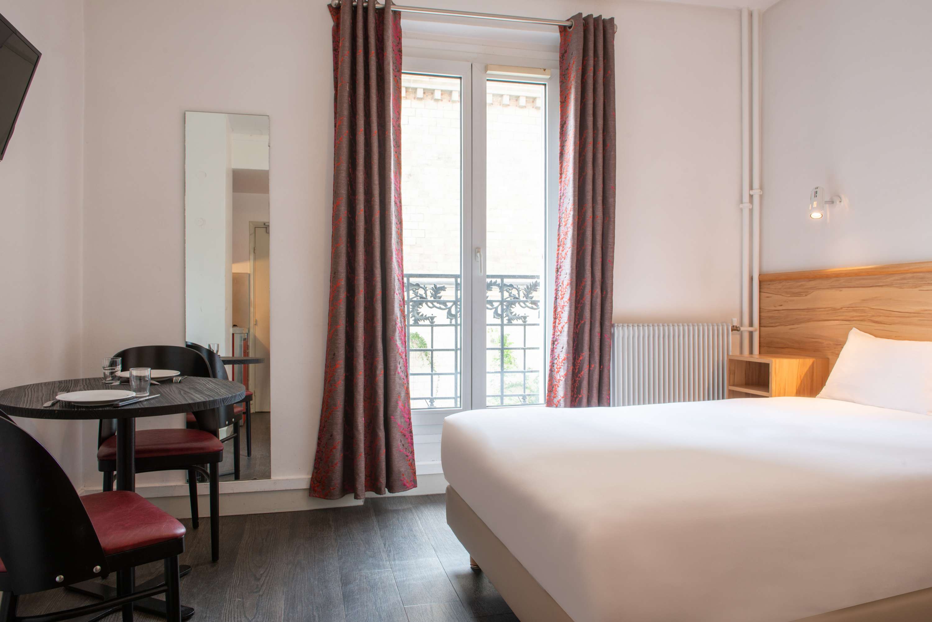 Image de studio de 15 m² avec balcon pour 2 personnes avec kitchenette rouge et acier avec frigidaire, table à manger, et micro-onde. Ce studio contient aussi une salle de bain blanche avec douche, lavabo, miroir et toilettes. Il y a un lit double et une télévision.