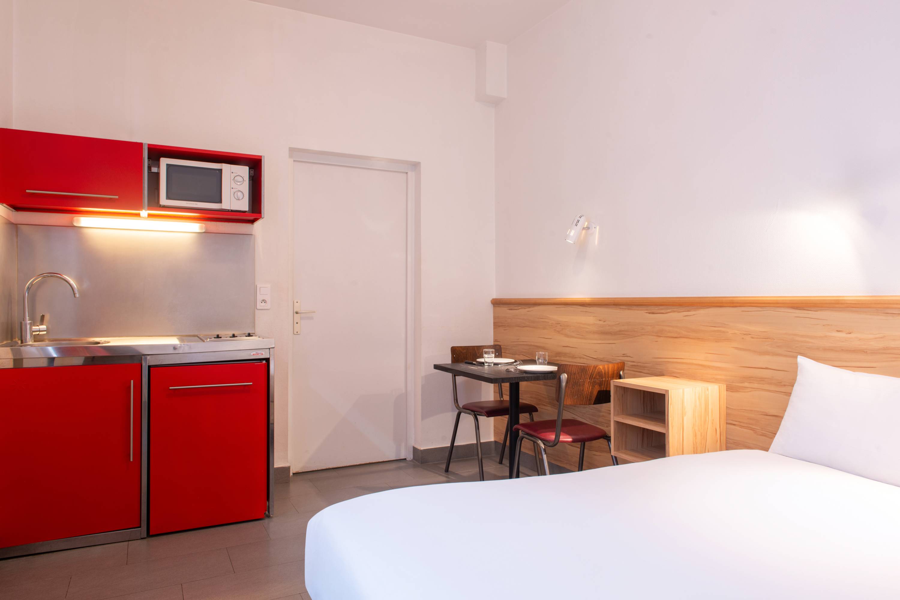 Image de studio de 12 m² vue sur cour pour 2 personnes avec kitchenette rouge et acier avec frigidaire, table à manger, et micro-onde. Ce studio contient aussi une salle de bain blanche avec douche, lavabo, miroir et toilettes. Il y a un lit double et une télévision.