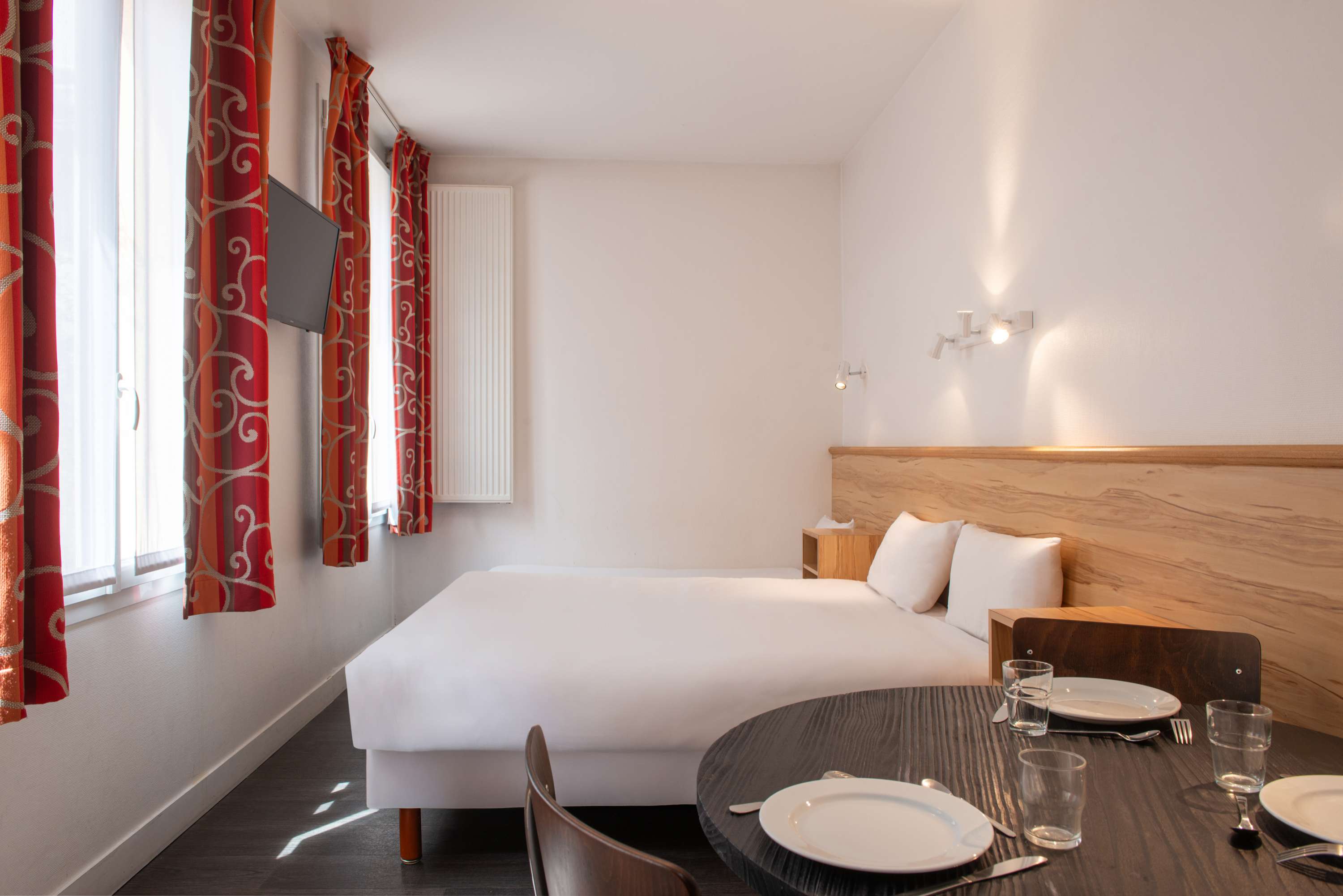 Image de studio de 15 m² pour 2 personnes avec kitchenette rouge et acier avec frigidaire, table à manger, et micro-onde. Ce studio contient aussi une salle de bain blanche avec douche, lavabo, miroir et toilettes. Il y a deux lits individuels et une télévision.
