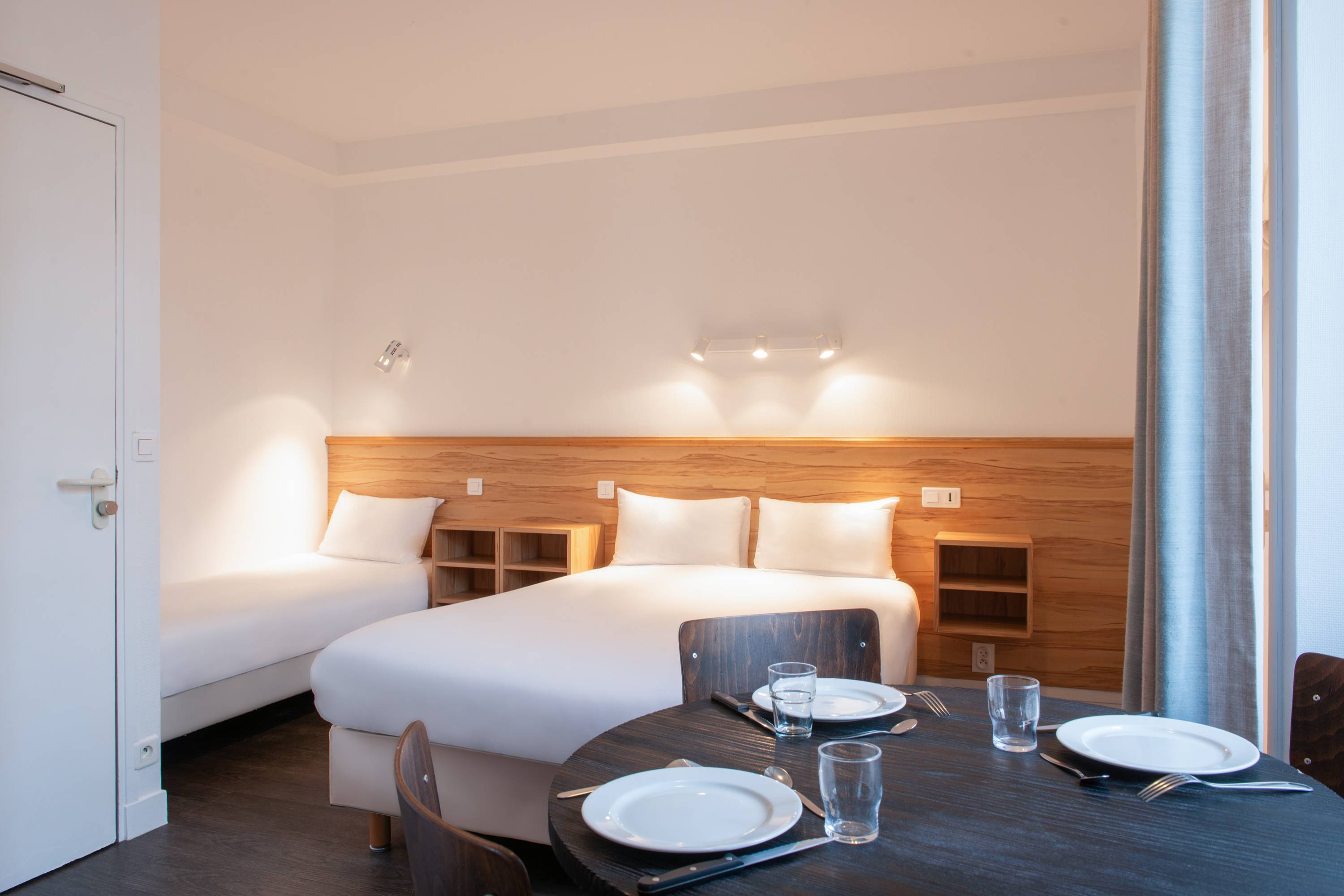 Image de studio de 19 m² pour 3 personnes avec kitchenette rouge et acier avec frigidaire, table à manger, et micro-onde. Ce studio contient aussi une salle de bain blanche avec douche, lavabo, miroir et toilettes. Il y a un lit double, un lit individuel et une télévision.