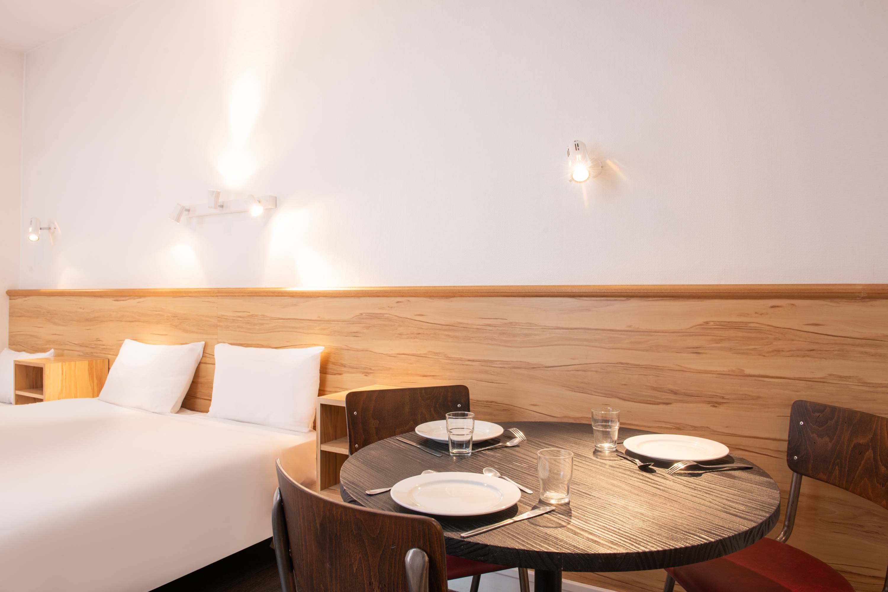 Image de studio de 19 m² vue sur cour pour 3 personnes avec kitchenette rouge et acier avec frigidaire, table à manger, et micro-onde. Ce studio contient aussi une salle de bain blanche avec douche, lavabo, miroir et toilettes. Il y a un lit double, un lit individuel et une télévision.