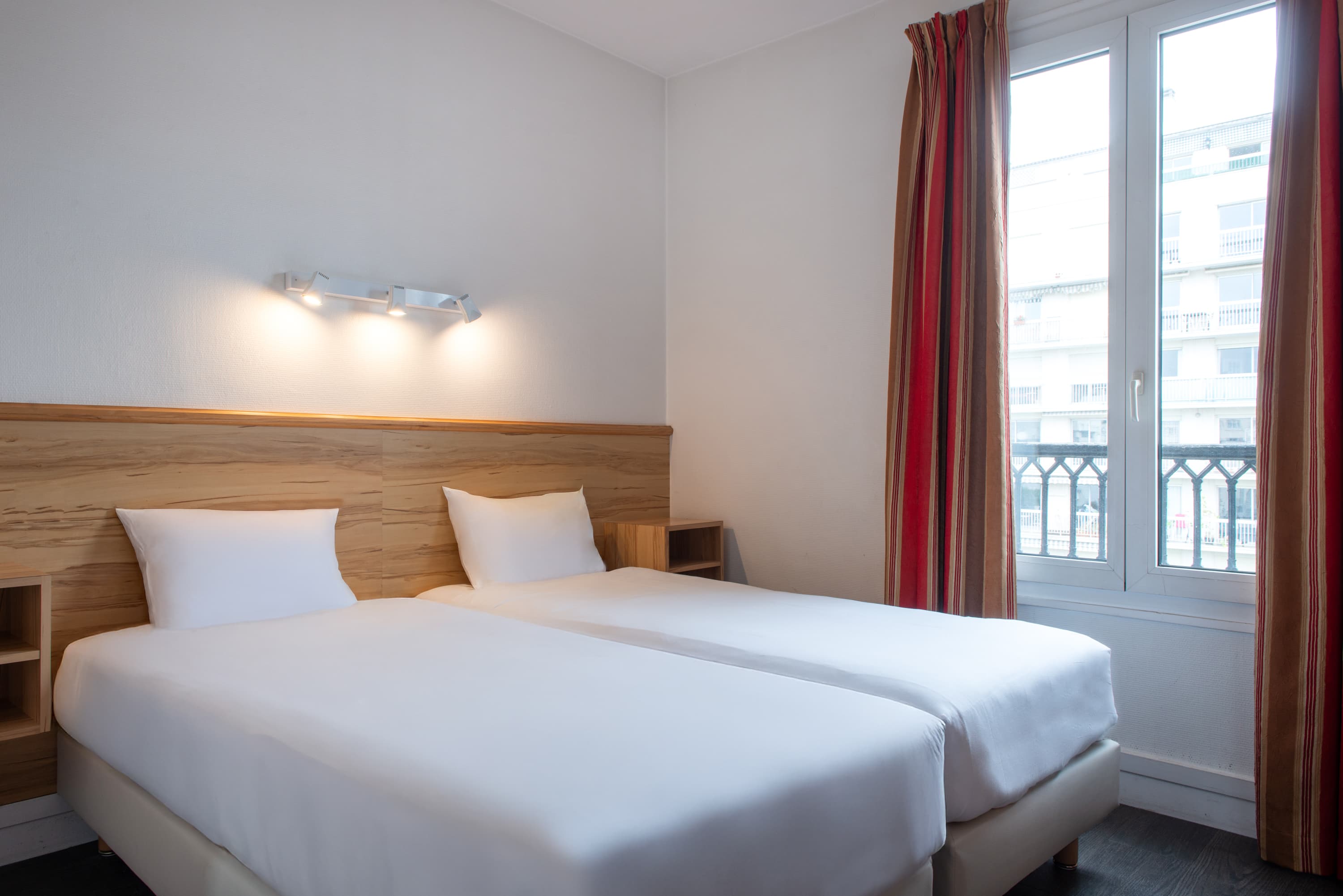 Image de studio de 15 m² pour 2 personnes avec kitchenette rouge et acier avec frigidaire, table à manger, et micro-onde. Ce studio contient aussi une salle de bain blanche avec douche, lavabo, miroir et toilettes. Il y a deux lits individuels et une télévision.