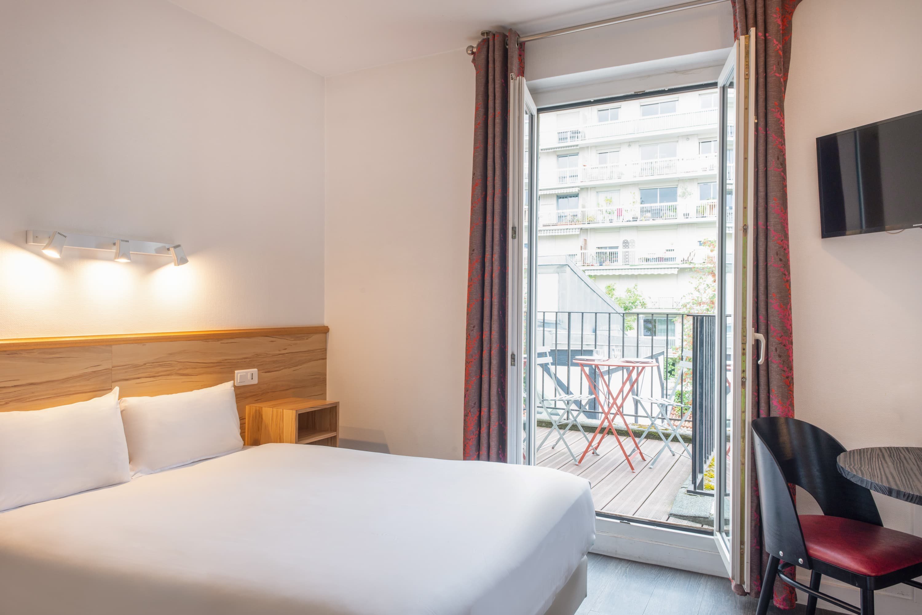 Image de studio de 15 m² avec terrasse pour 2 personnes avec kitchenette rouge et acier avec frigidaire, table à manger, et micro-onde. Ce studio contient aussi une salle de bain blanche avec douche, lavabo, miroir et toilettes. Il y a un lit double et une télévision.