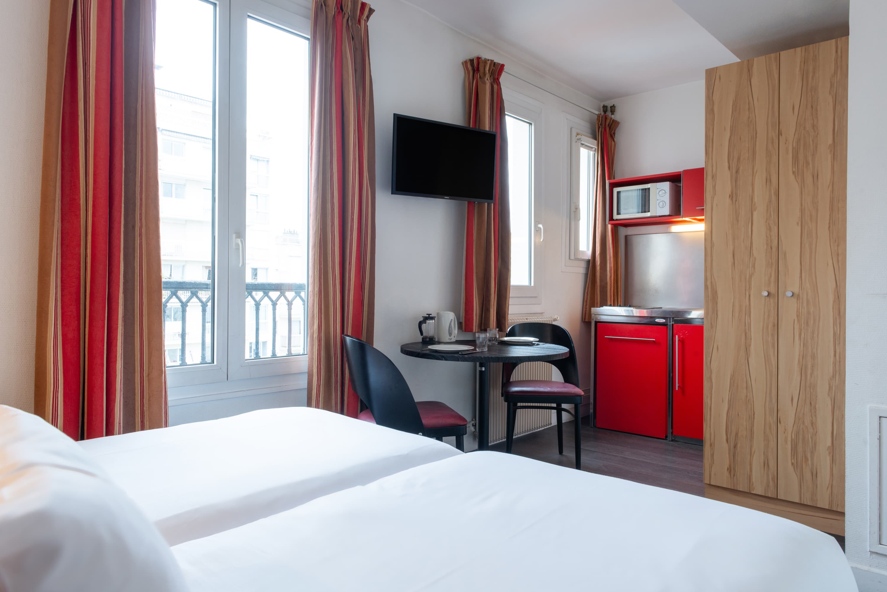 Image de studio de 15 m² pour 2 personnes avec kitchenette rouge et acier avec frigidaire, table à manger, et micro-onde. Ce studio contient aussi une salle de bain blanche avec douche, lavabo, miroir et toilettes. Il y a deux lits individuels et une télévision.