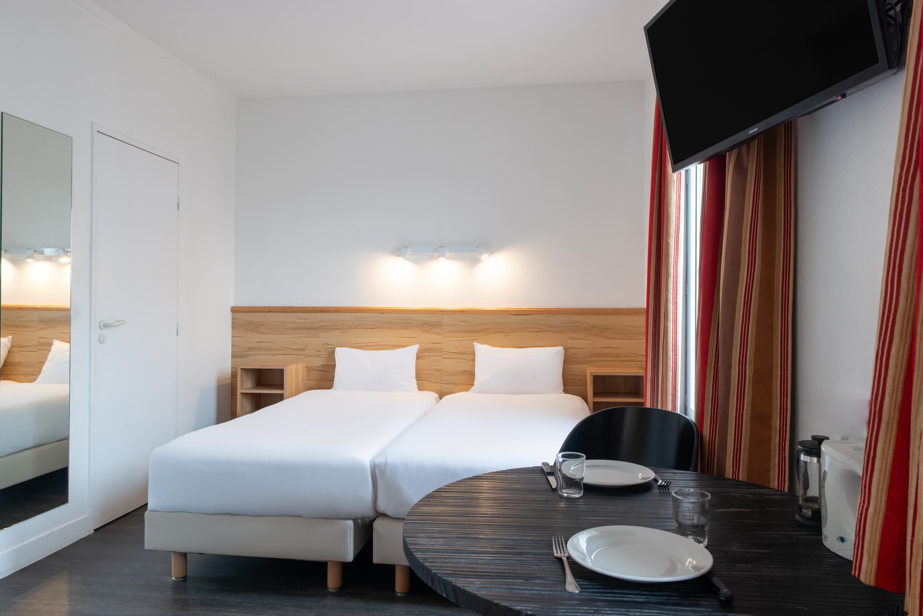 Image de studio de 15 m² pour 2 personnes avec kitchenette rouge et acier avec frigidaire, table à manger, et micro-onde. Ce studio contient aussi une salle de bain blanche avec douche, lavabo, miroir et toilettes. Il y a deux lits individuels et une télévision.