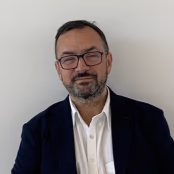 Smiling middle-aged man with glasses, beard, wearing a white shirt and dark blazer against a white wall.