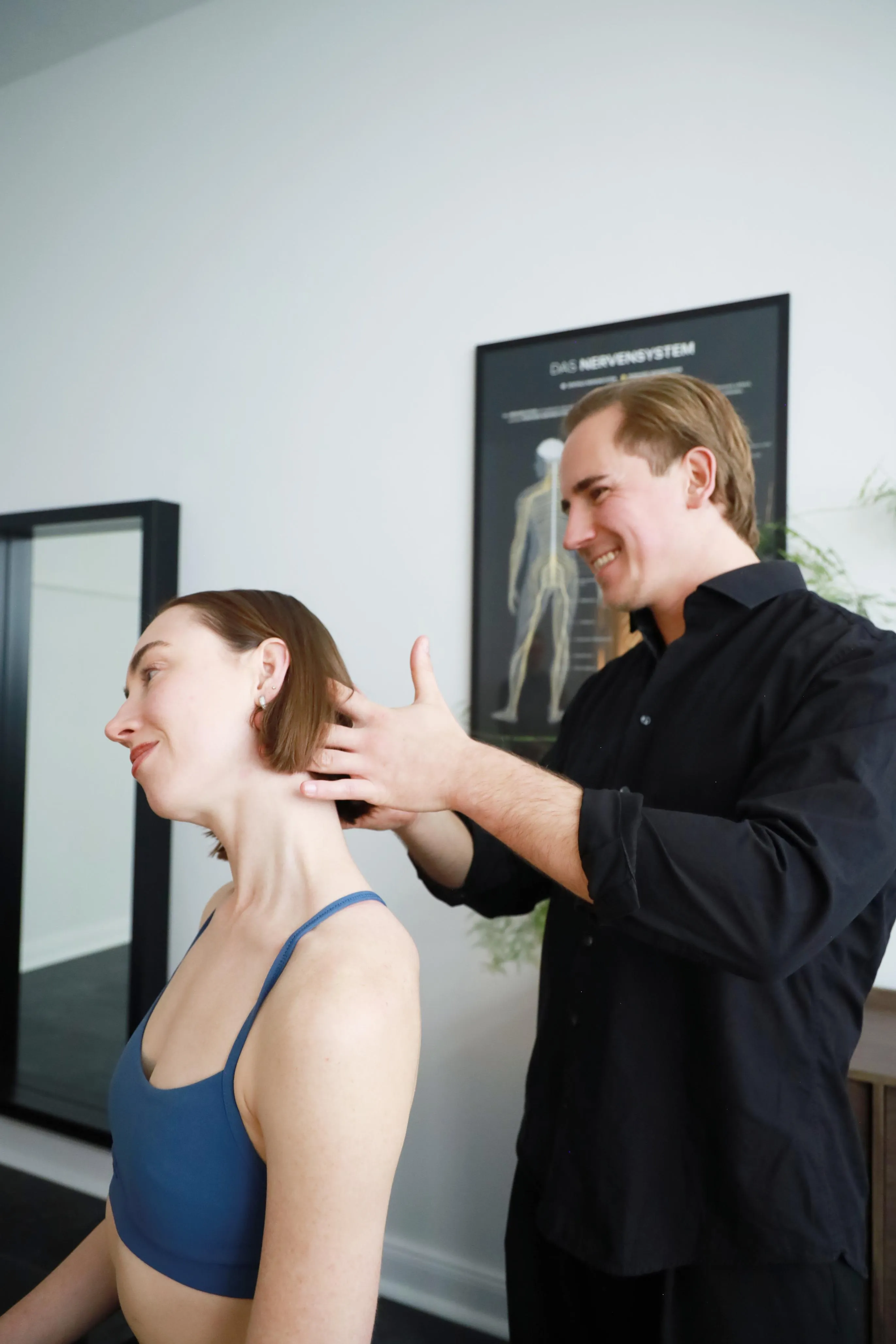 Ein Mann in schwarzem Hemd massiert sanft den Nacken einer in Blau gekleideten Frau in einem Therapieraum.
