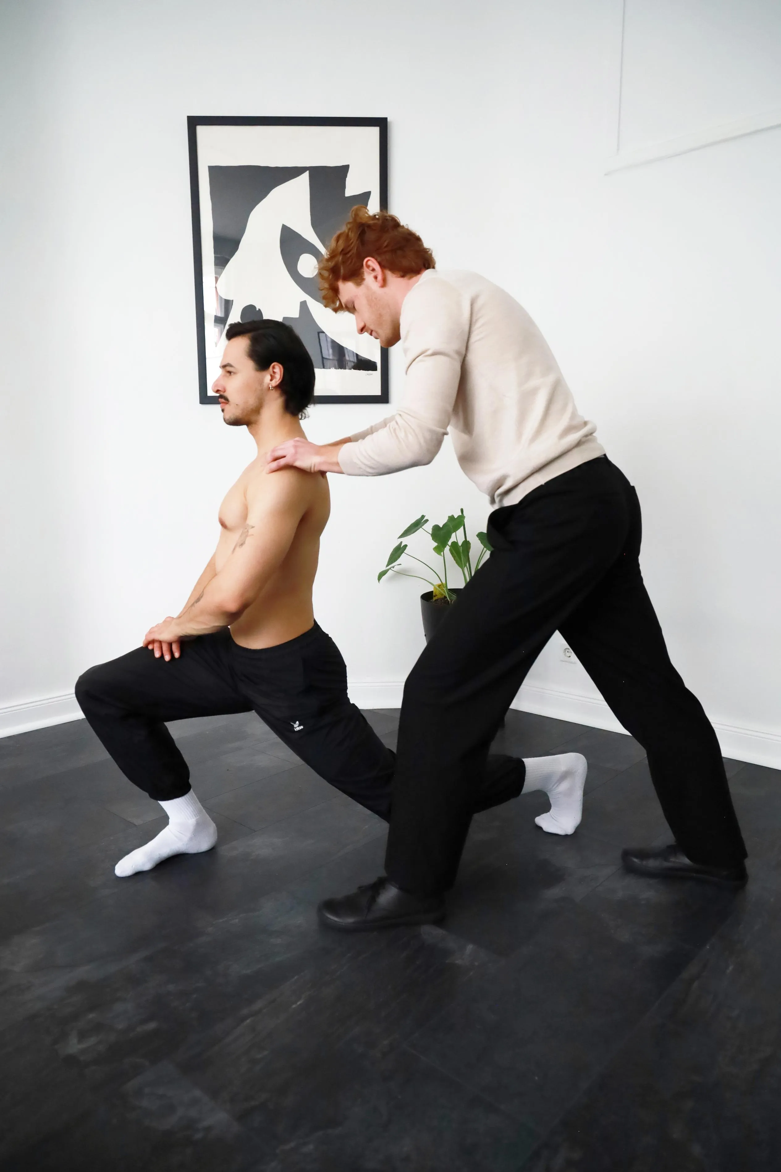 Ein Mann mit hellem Pullover und schwarzer Hose dehnt die Schultern eines anderen Mannes mit schwarzer Jogginghose und weißen Socken, der in einer tiefen Ausfallschrittposition steht.