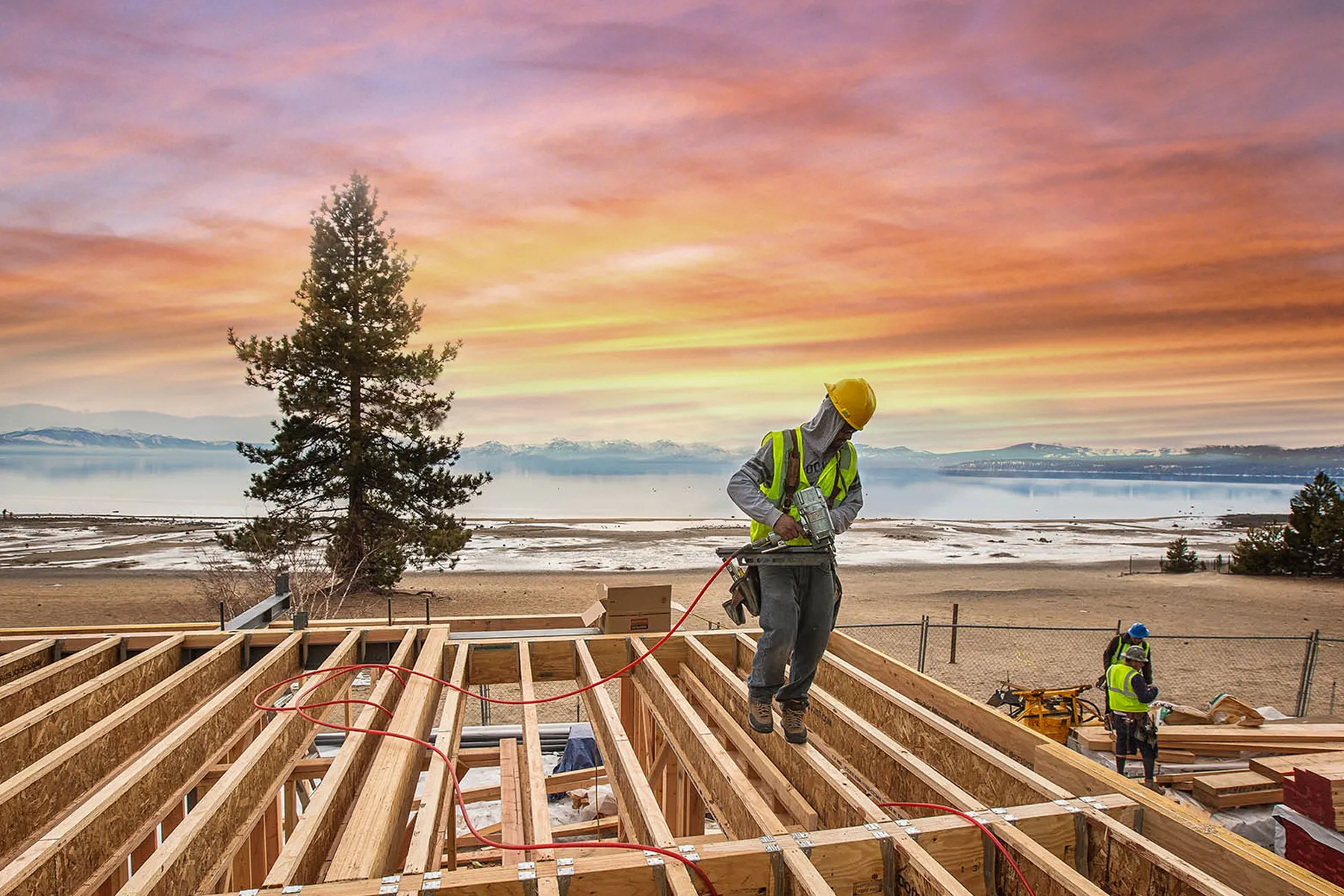 Construction worker in a yellow hard hat and vest using a nail gun while jumping between wooden beams with a serene lake and colorful sunset in the background.