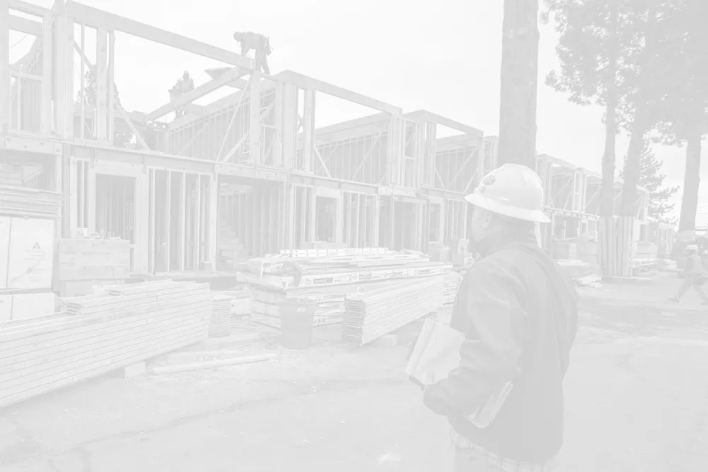 Construction worker wearing a white hard hat and holding blueprints, overseeing timber-framed buildings under construction.