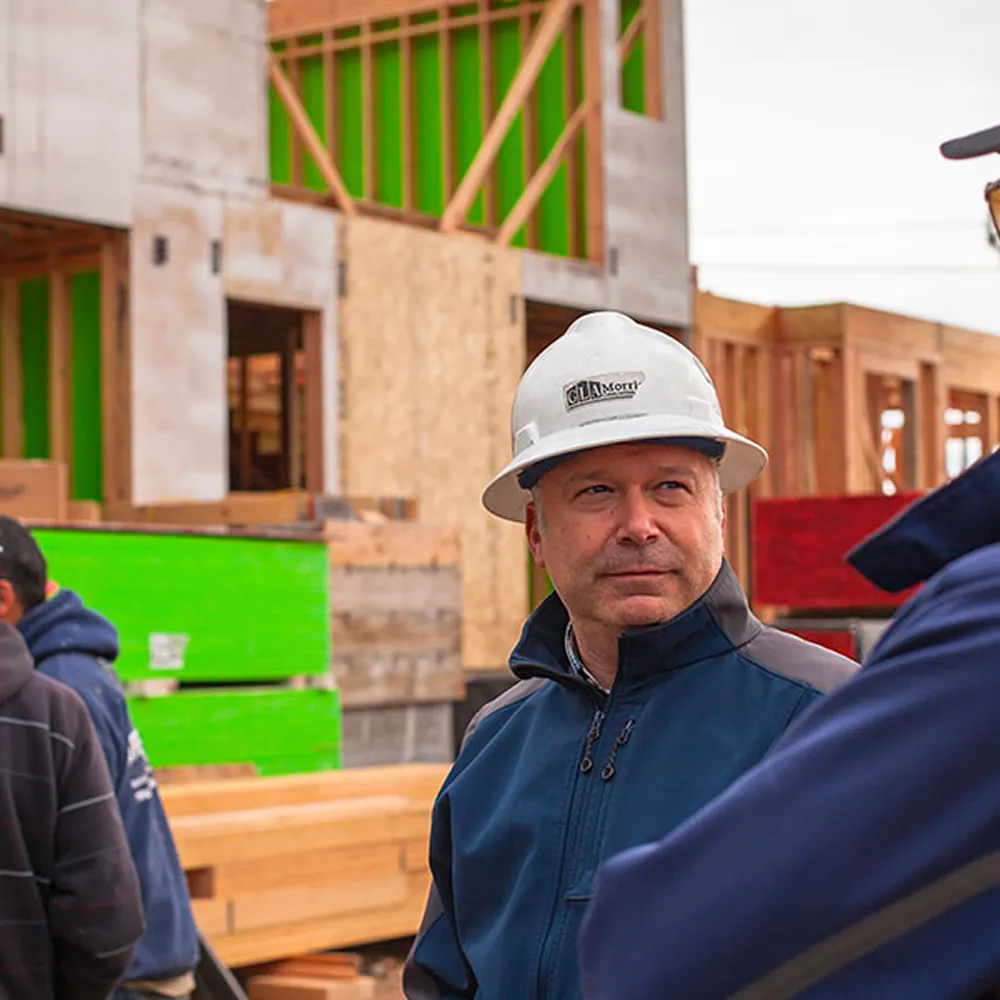 Construction worker wearing a white hard hat and navy jacket at a building site with wooden frames and green panels in the background.