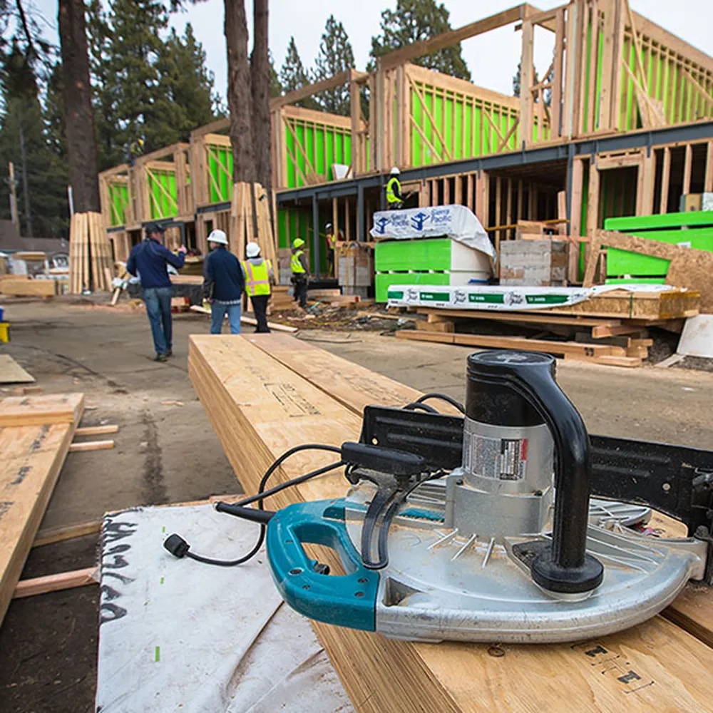 Circular saw resting on wooden planks at a residential construction site with workers and framed structures in the background.