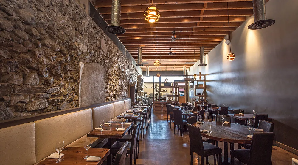 Dining area with wooden tables, leather chairs, stone wall on the left, and multiple hanging lights from a wooden beam ceiling.